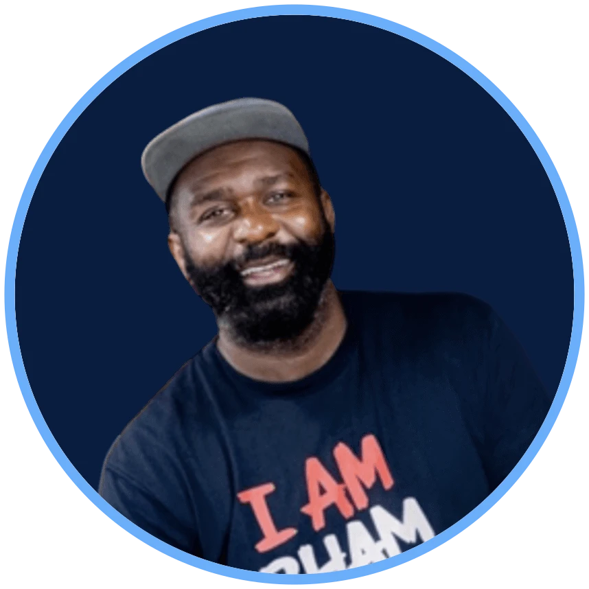Portrait of a man with a beard wearing a gray cap and a black T-shirt that says 'I AM BHAM,' smiling against a blue background.