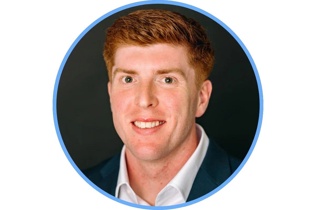 A headshot of a man with red hair, wearing a white shirt and dark suit jacket, smiling against a dark background.